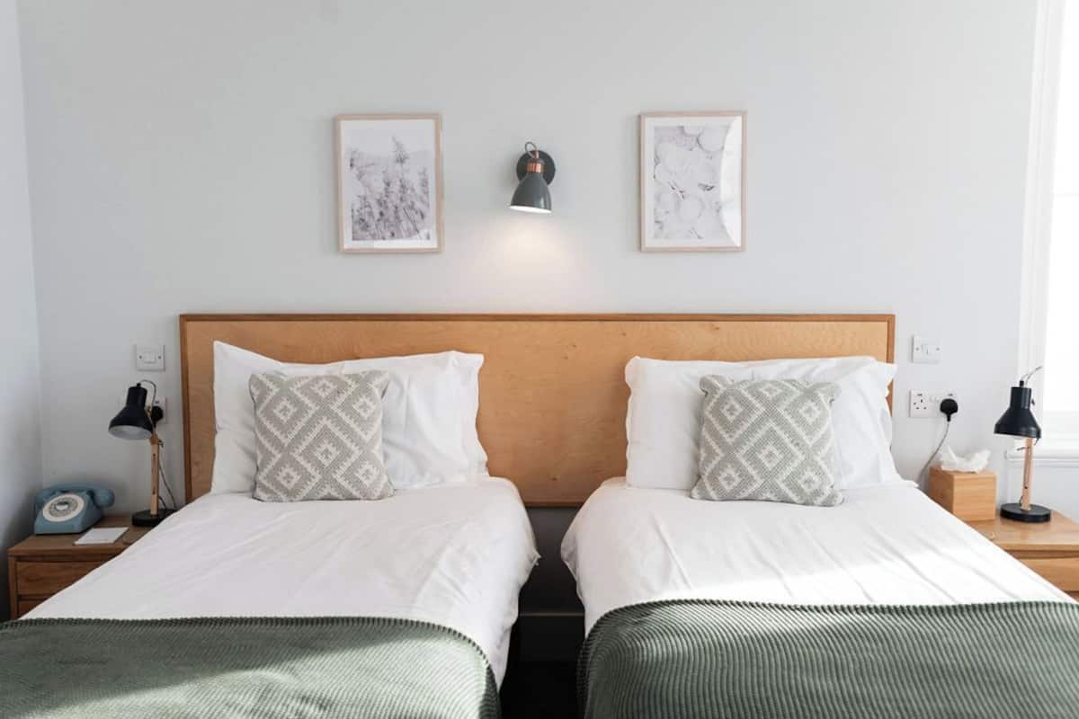 Een tweepersoonskamer met twee bedden, elk voorzien van een houten hoofdbord, wit beddengoed en gestreepte kussens. Er staan nachtkastjes met lampen bij de bedden, een wandlamp en boven de bedden hangt kunst in lijsten.