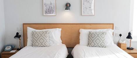Habitación doble con dos camas, cada una con cabecero de madera, ropa de cama blanca y almohadas estampadas. Hay mesitas de noche con lámparas, una luz de pared y cuadros colgados sobre las camas.