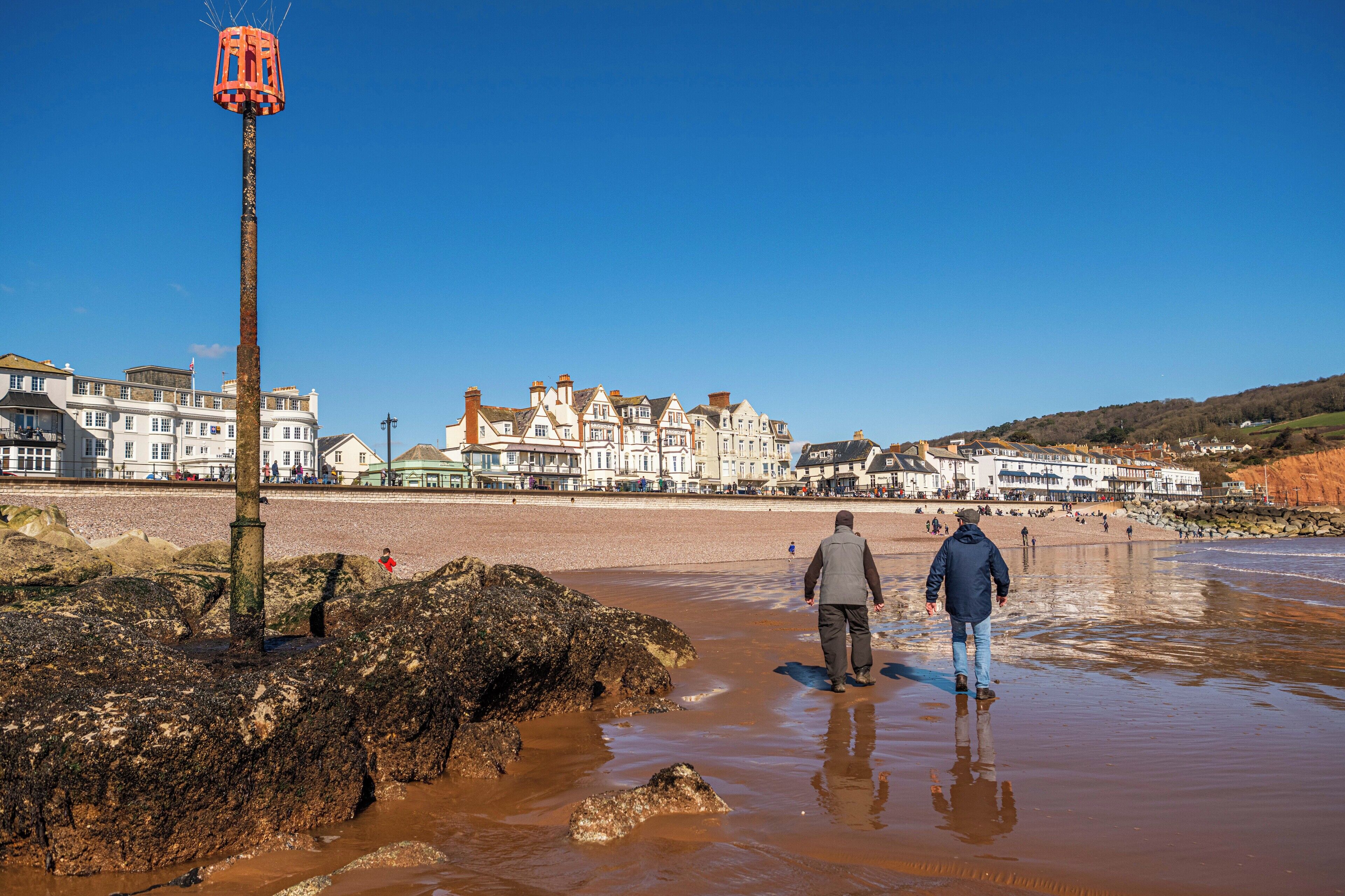 A coastal town with buildings along the shore, a rocky area with a lighthouse, and people walking on the beach.