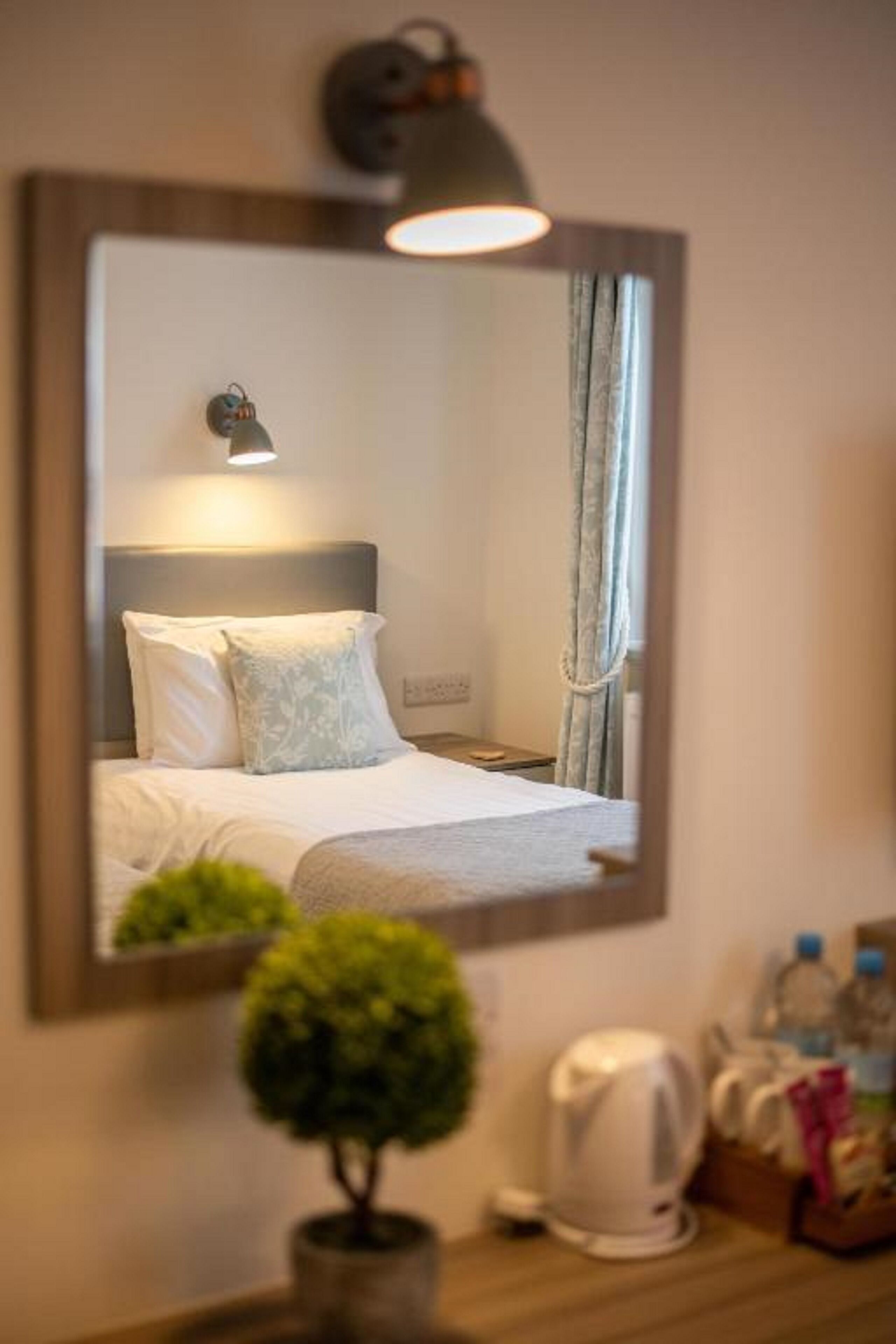A hotel room with a bed, a bedside table with a kettle, a small potted plant, and a mirror reflecting the room.