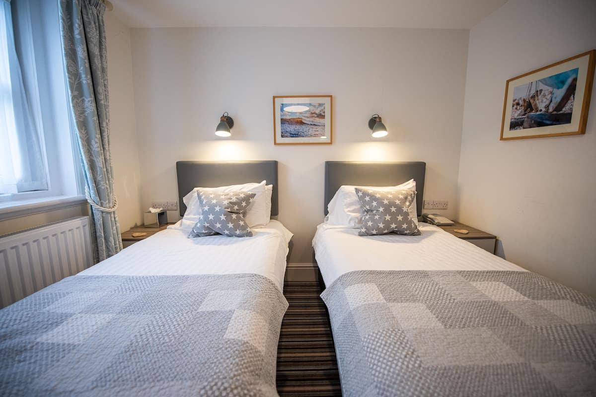 Hotelkamer met twee bedden, een raam met gordijnen, een lijst met een foto aan de muur en een nachtkastje met een telefoon.