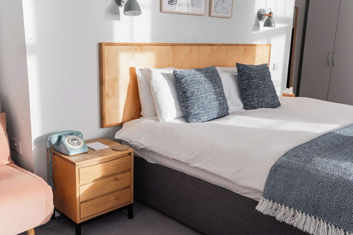 Een netjes opgemaakt slaapkamer met een houten hoofdbord, een bed met witte en gemêleerde kussens, een nachtkastje met een draaischijf telefoon, en een nachtkastje.