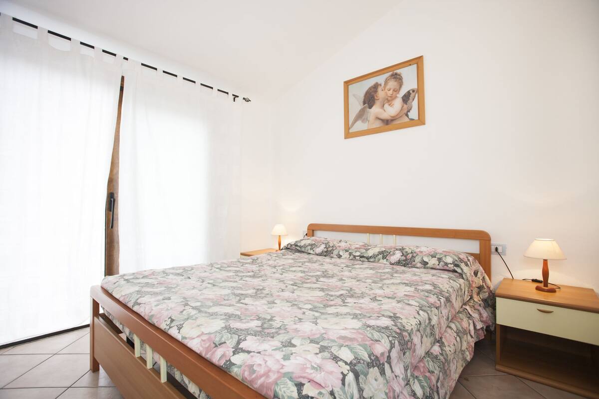 A bedroom with a wooden bed, floral bedding, a nightstand with a lamp, a wall-mounted picture, and a window with sheer curtains.