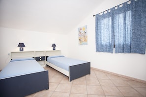 Quarto com duas camas de solteiro, uma escrivaninha com duas luminárias e uma janela com cortinas azuis.