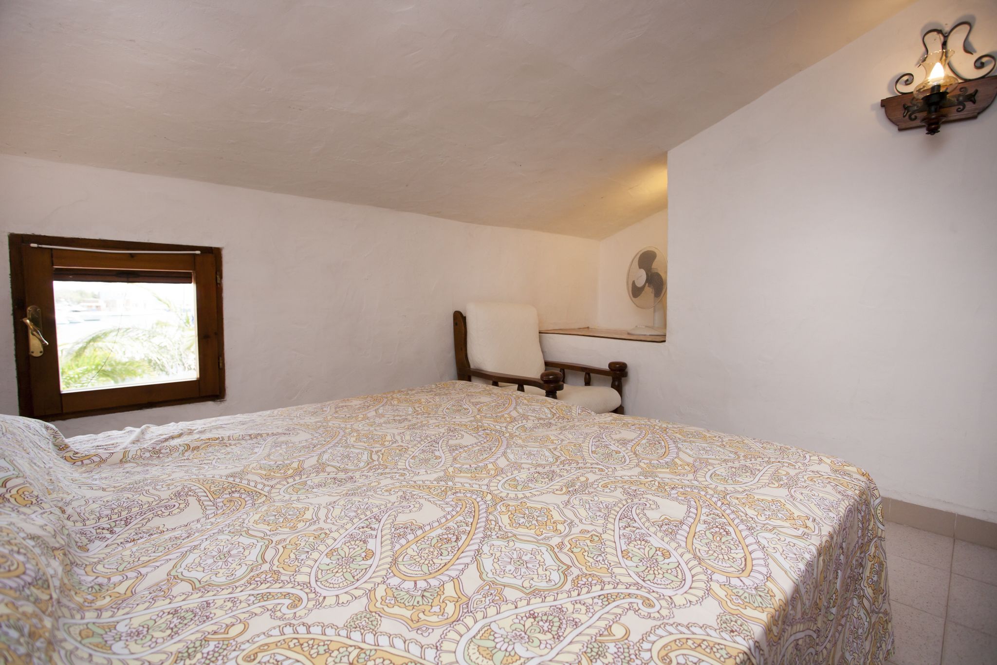 A bedroom with a bed, a chair, a window, and a wall-mounted light fixture.
