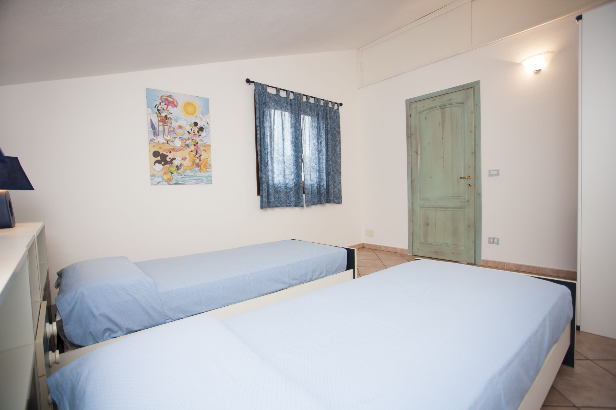 Quarto com duas camas de solteiro, uma janela com cortinas, uma porta e uma parede com um cartaz.
