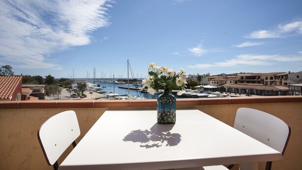 Un balcón con una mesa y sillas blancas, un jarrón con flores, y vista a un puerto deportivo con barcos y edificios.