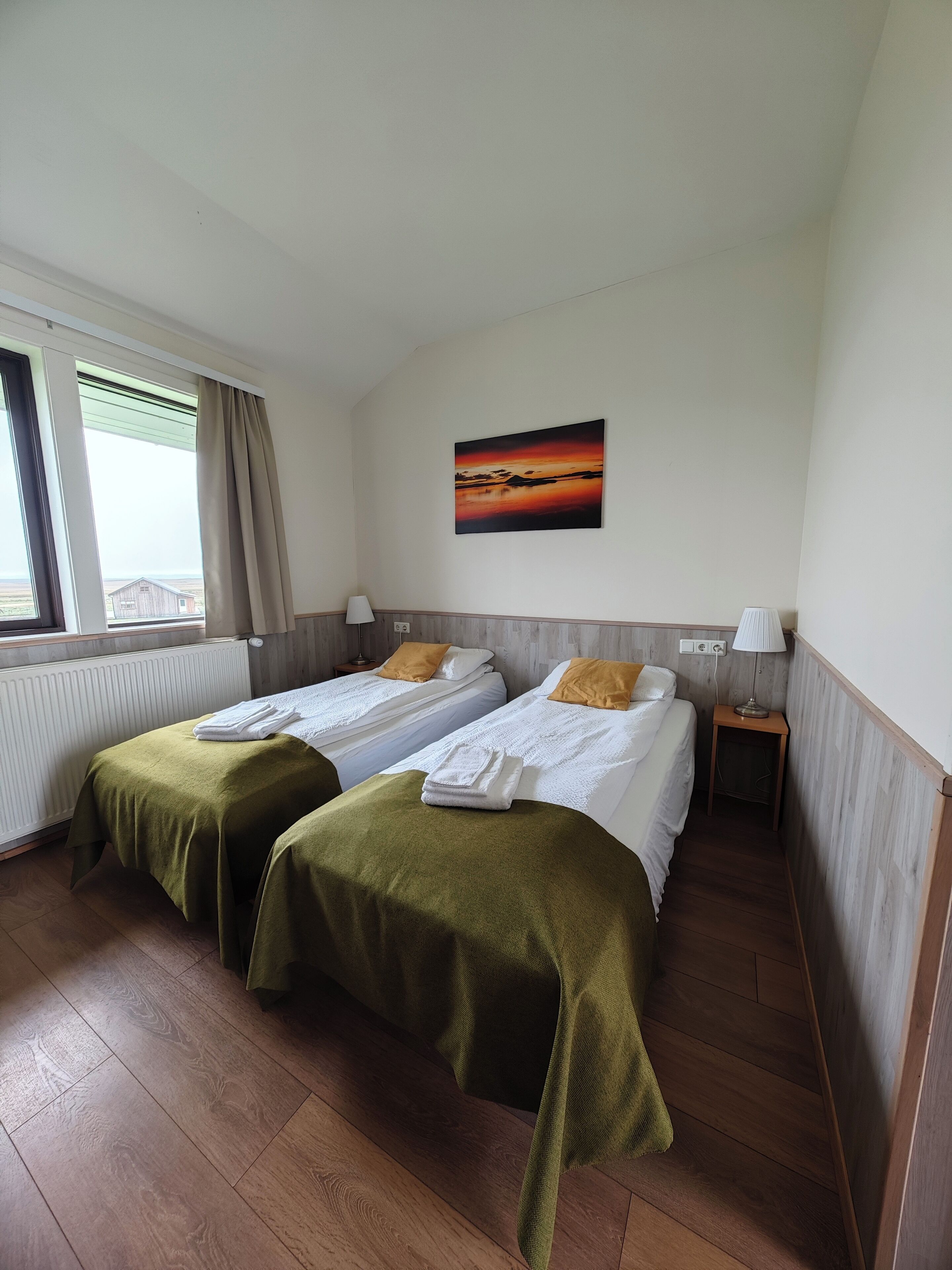 Een hotelkamer met twee bedden, een schilderij van een zonsondergang, een raam met gordijnen en een radiator.
