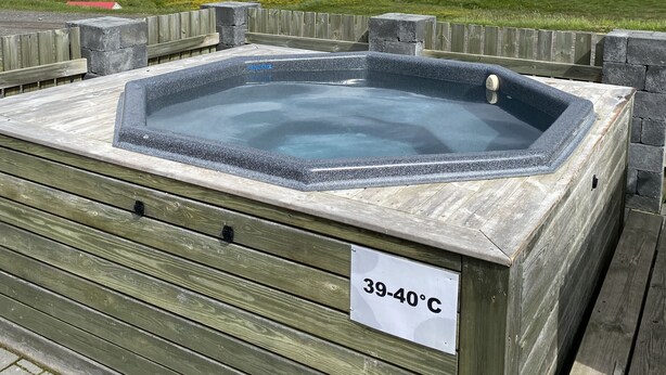 A hot tub with a sign indicating a temperature range of 39-40°C, surrounded by a wooden deck and a grassy field with mountains in the background.