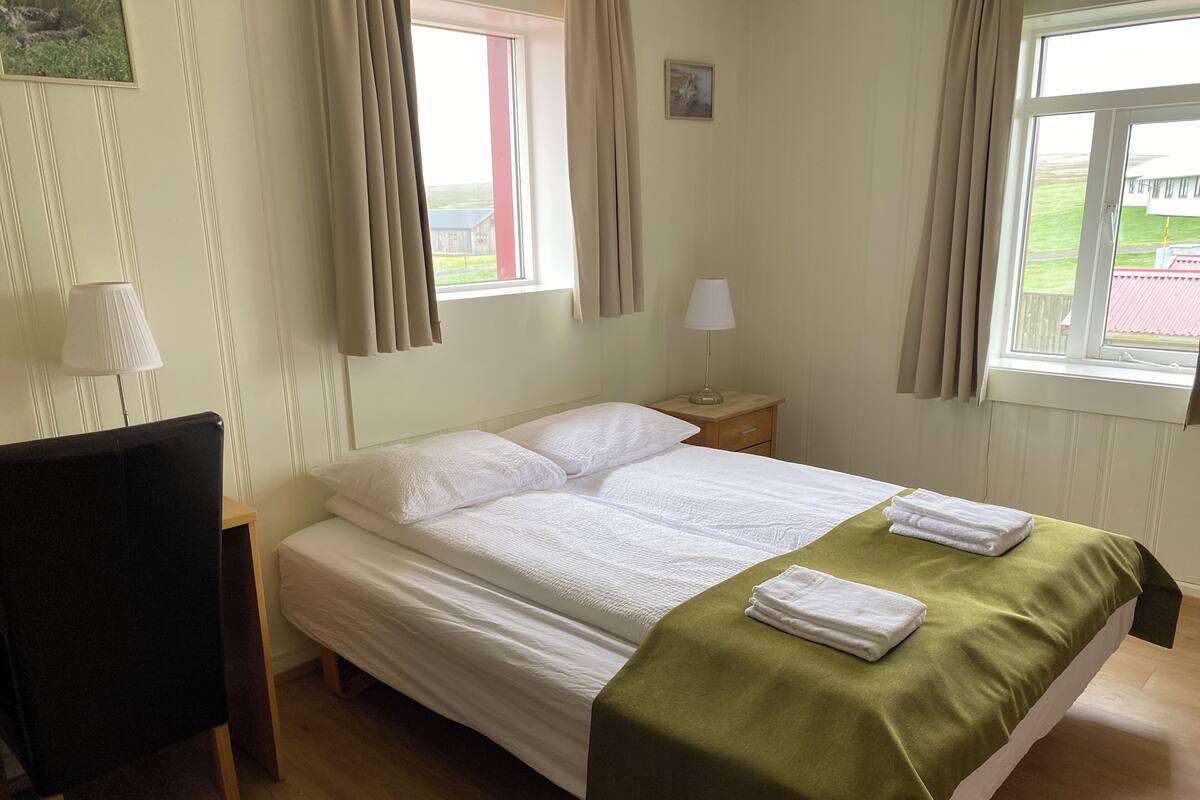 Een netjes opgemaakt bed met witte lakens en een groene plaid, een nachtkastje met een lamp, en een stoel links.