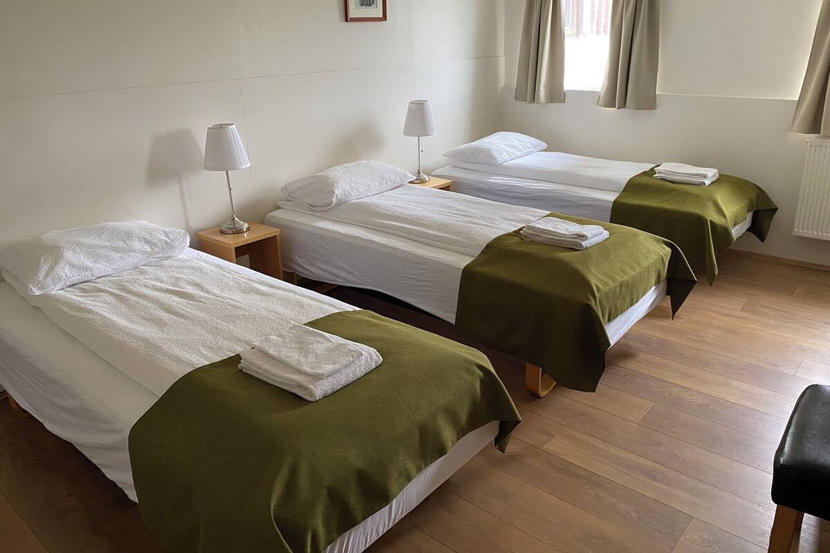 Een hotelkamer met drie enkele bedden, elk met een groene sprei en witte lakens. Twee nachtlampjes, een inlijsting foto aan de muur en een raam met gordijnen.