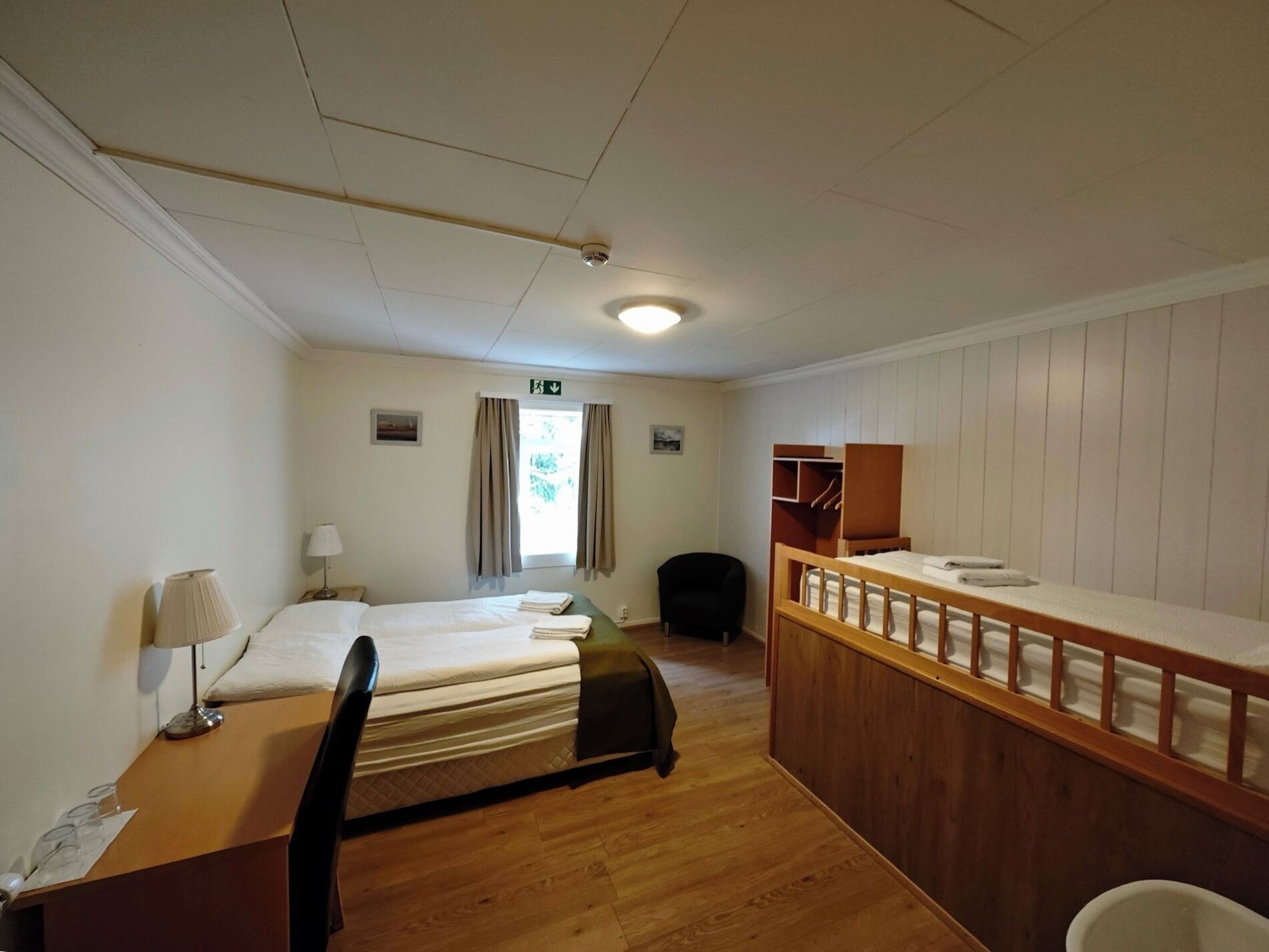 A hotel room with two single beds, a wooden desk, a chair, a small table with a lamp, a wooden bench, and a window with curtains.