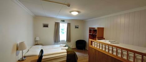 A hotel room with two single beds, a wooden desk, a chair, a small table with a lamp, a wooden bench, and a window with curtains.