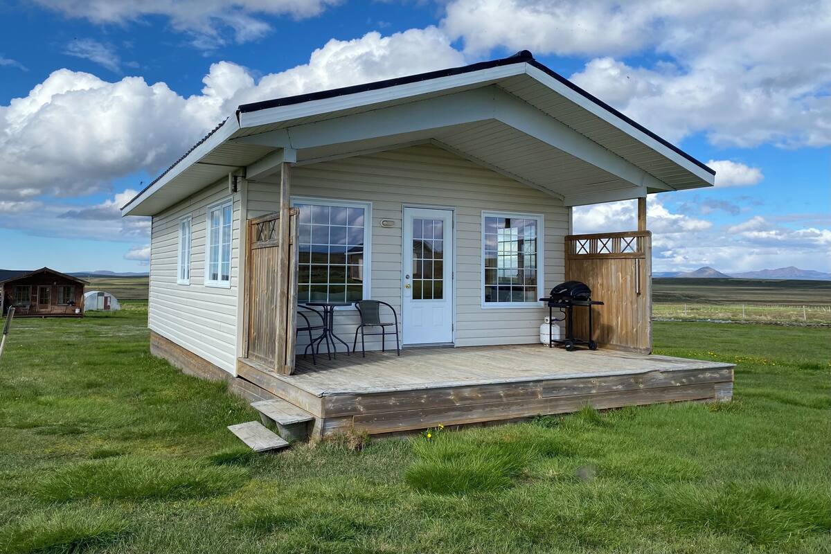 Een kleine, gelijkvloerse blokhut met een veranda, een barbecue en een raam met uitzicht op een grasveld.
