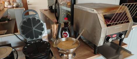 A kitchen with a waffle maker, a hot plate, a bowl with a spoon, and a hot grill.