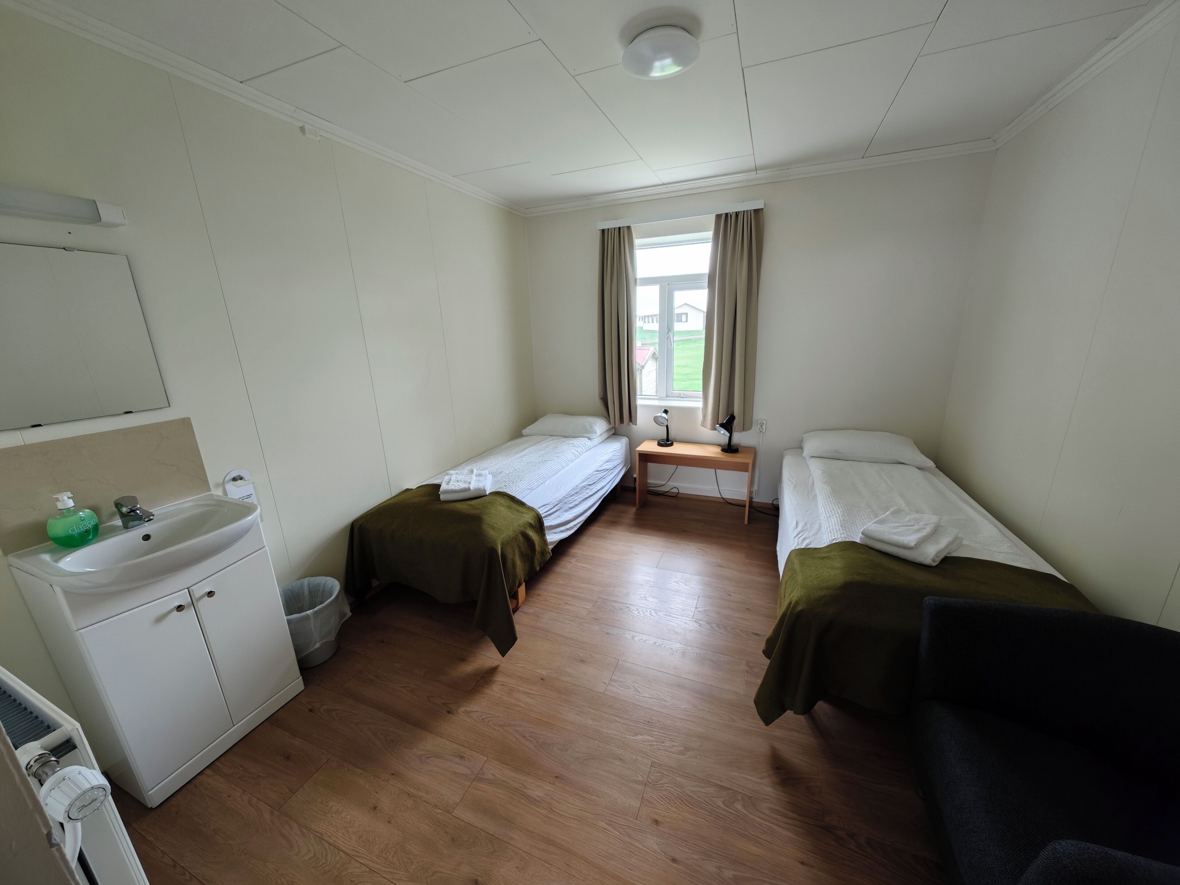 A small, clean room with two single beds, a wooden floor, a sink, and a window with curtains.