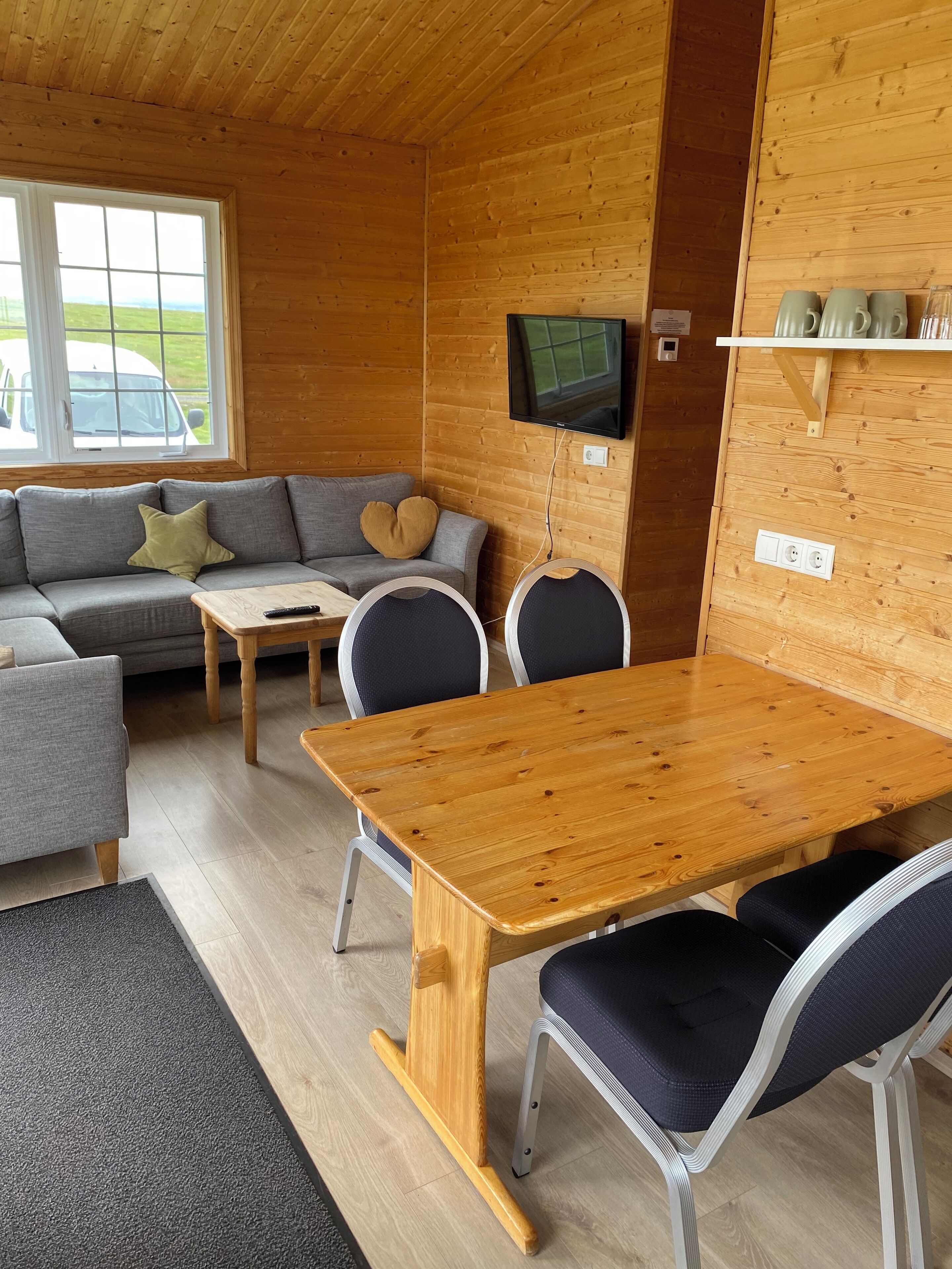 A cozy cabin interior with a wooden dining table, grey sofa, and a TV mounted on the wall.