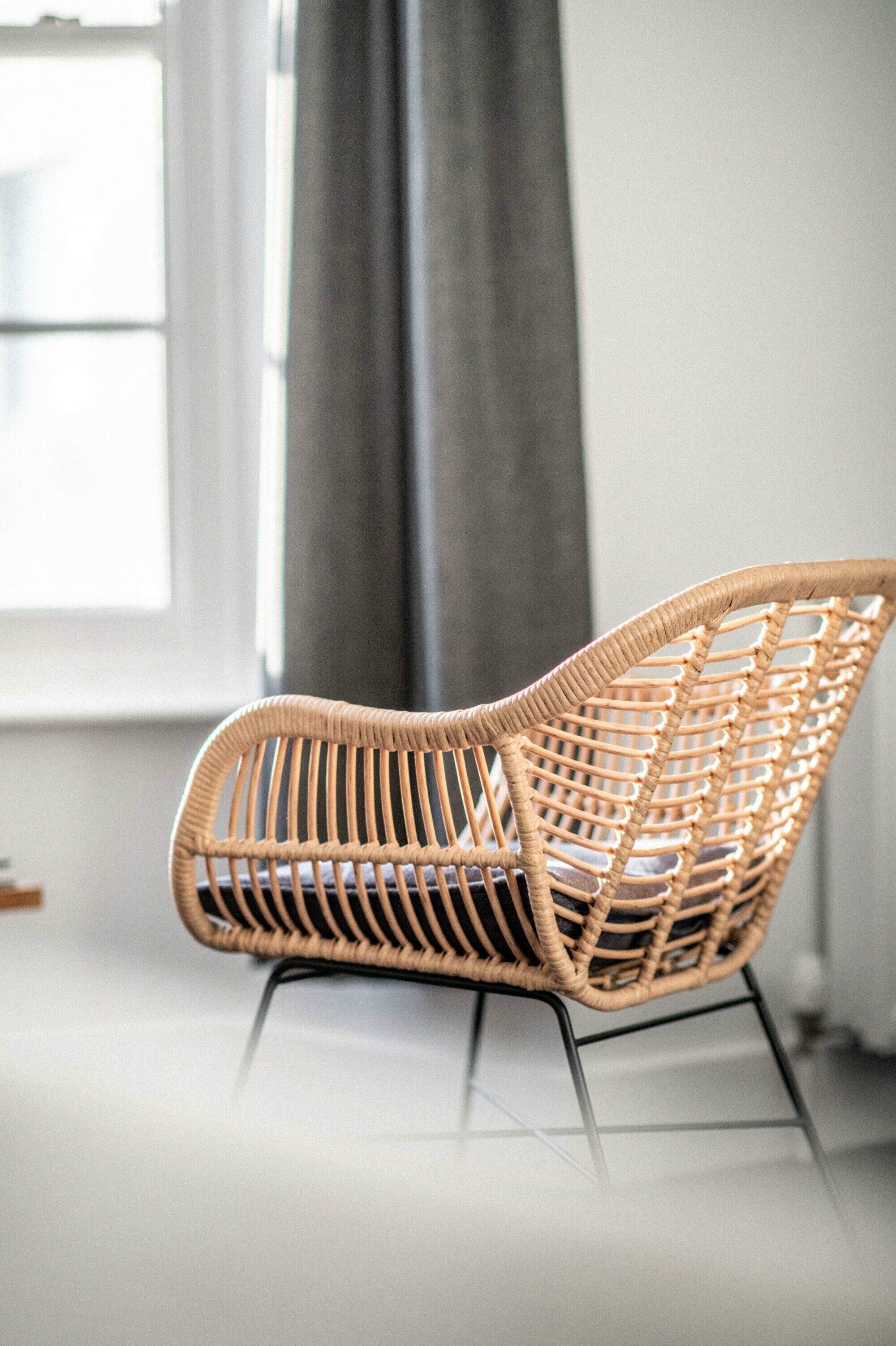 A wicker chair with a metal frame, positioned in a room with a window and a grey curtain.