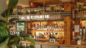 Zona de bar con estanterías llenas de botellas y vasos, una barra con grifo y un cartel que indica los precios de las bebidas.