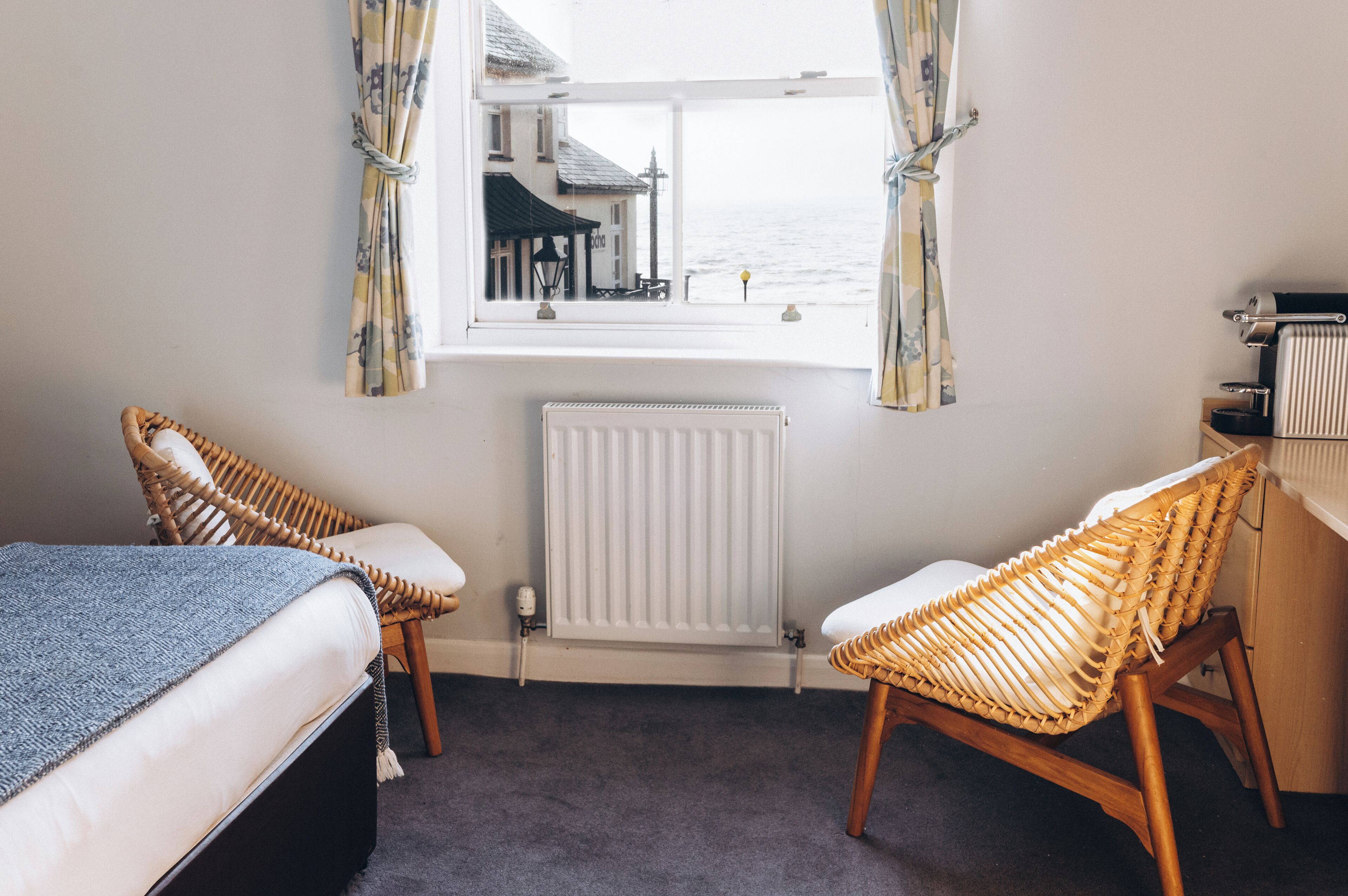 A room with two wicker chairs, a bed, a radiator, and a window overlooking a building and the sea.