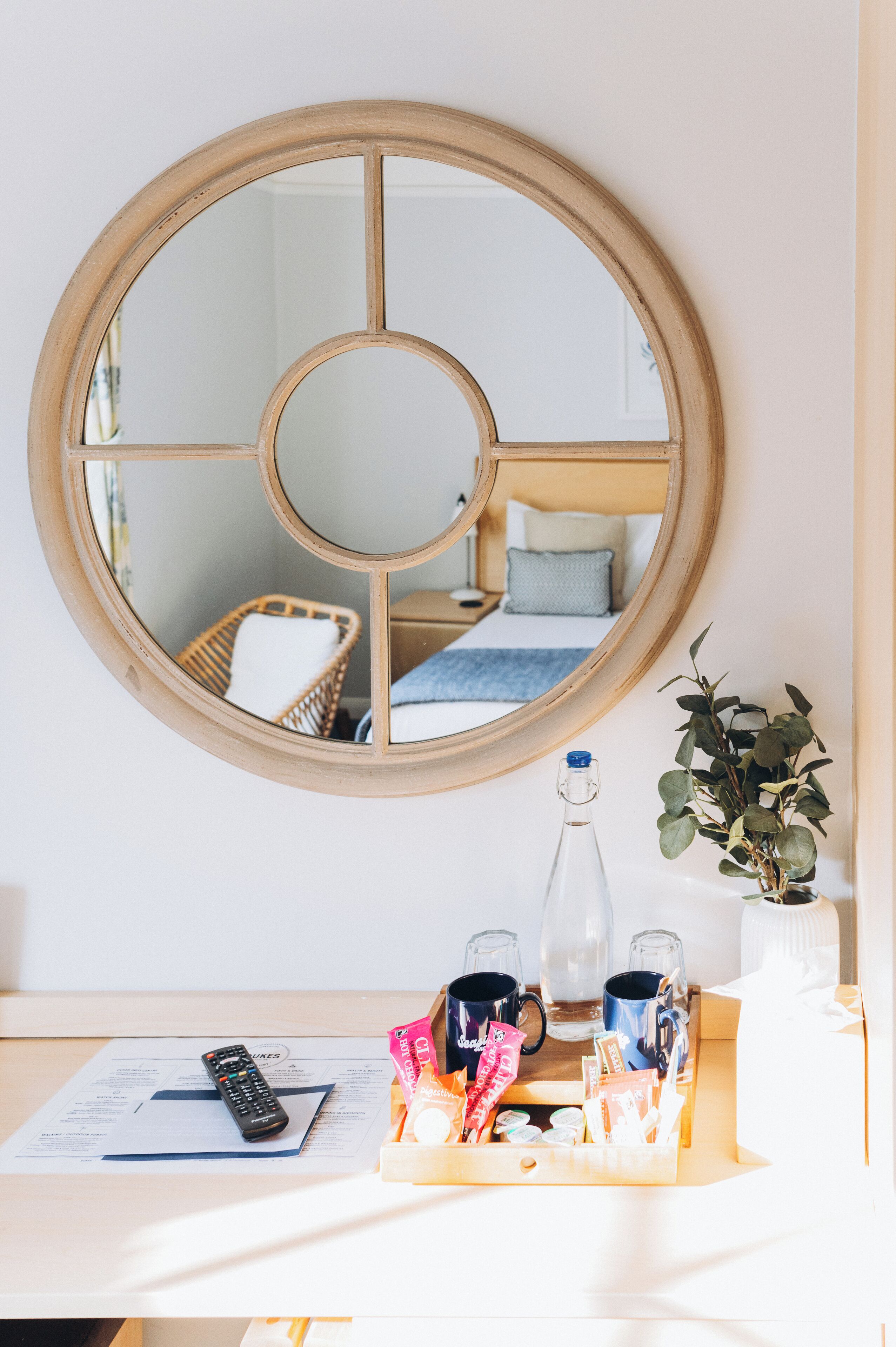 A round mirror with a wooden frame, a bedside table with a remote control, two mugs, a bottle, and a tray with snacks and a plant.