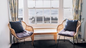 Dos sillas de mimbre con cojines, un banco de madera y un ventanal con vista al mar.