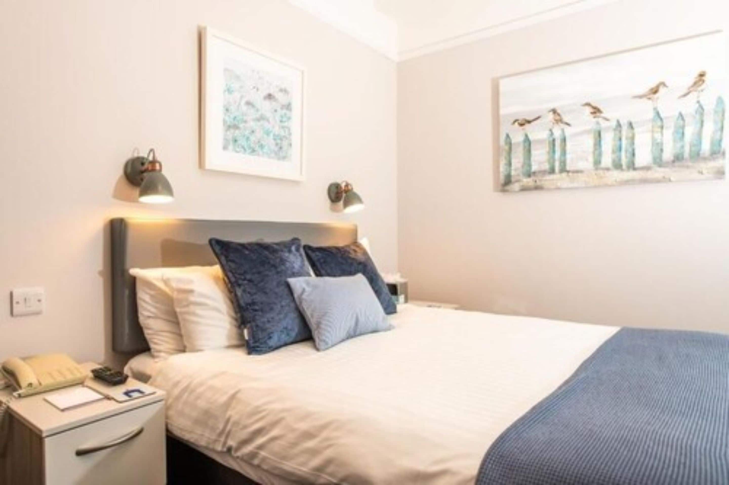 Een net opgemaakt bed met een blauw-witte dekbedovertrek, een bedlampje en een klein tafeltje met een telefoon en papieren.