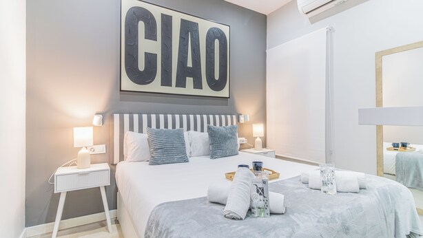 Une chambre d’hôtel moderne avec un grand lit, des tables de chevet avec des lampes, un climatiseur fixé au mur et une œuvre d’art encadrée sur laquelle figure le mot « CIAO ».