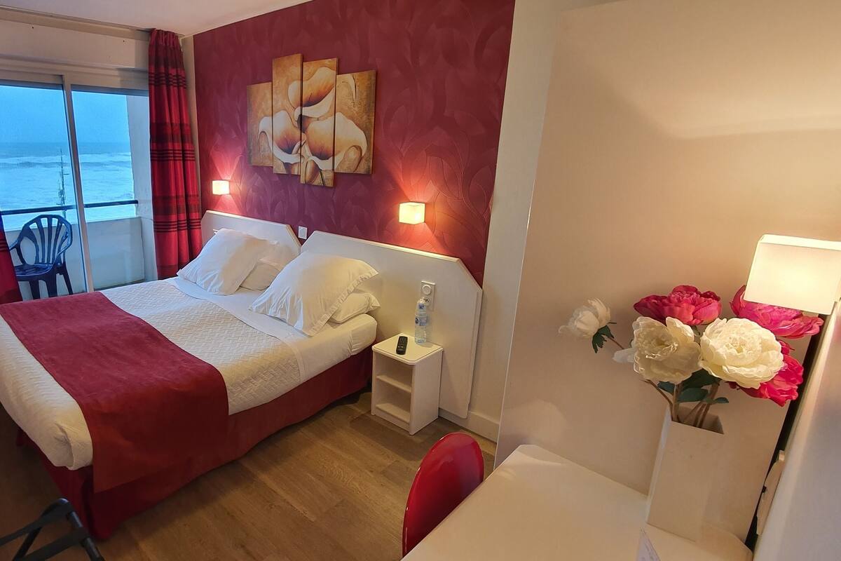 Habitación de hotel con cama, mesita de noche, silla, escritorio, ventana con vista al mar y un cuadro en la pared.