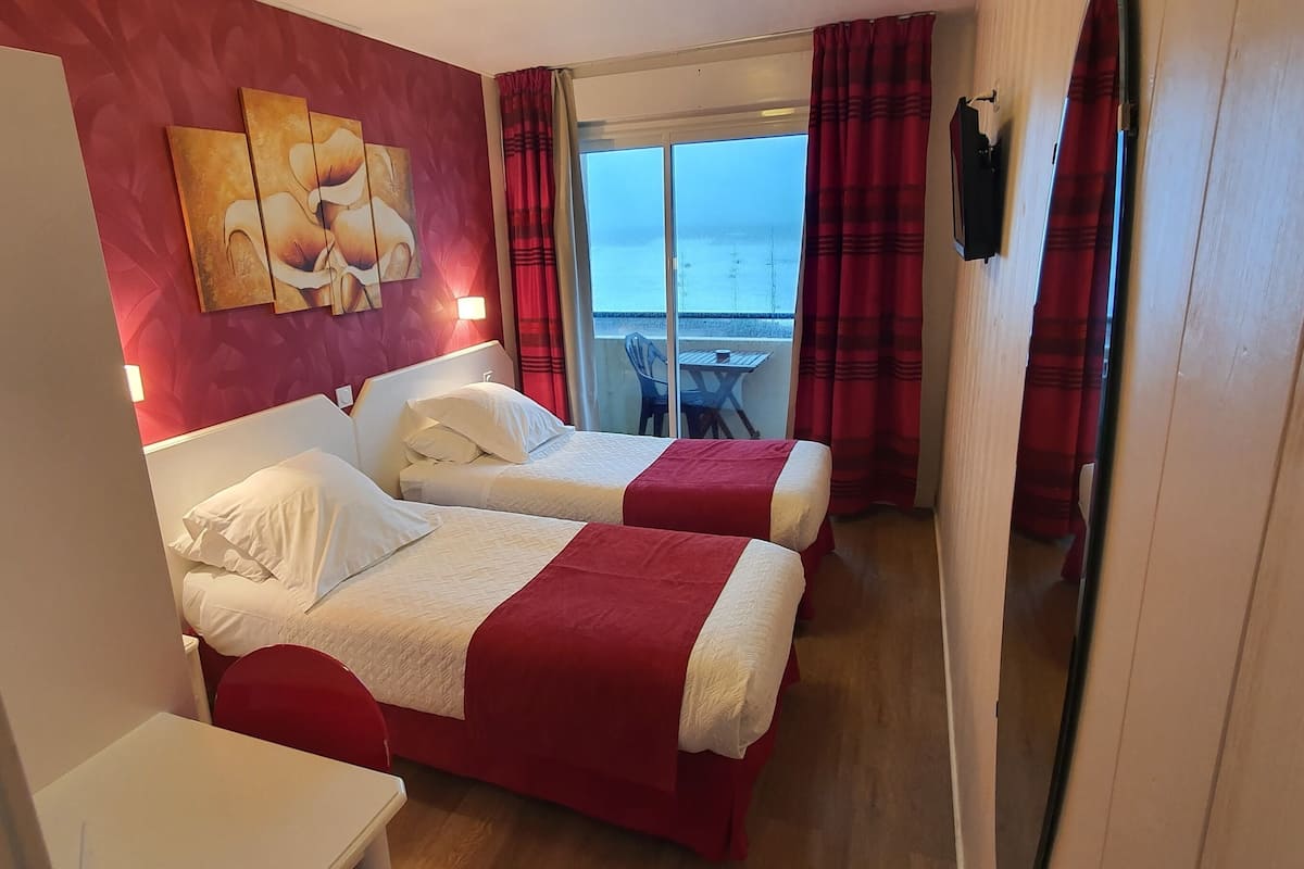 Habitación de hotel con dos camas, una decoración en tonos rojo y blanco, un cuadro en la pared y vista al mar a través de una puerta corredera.