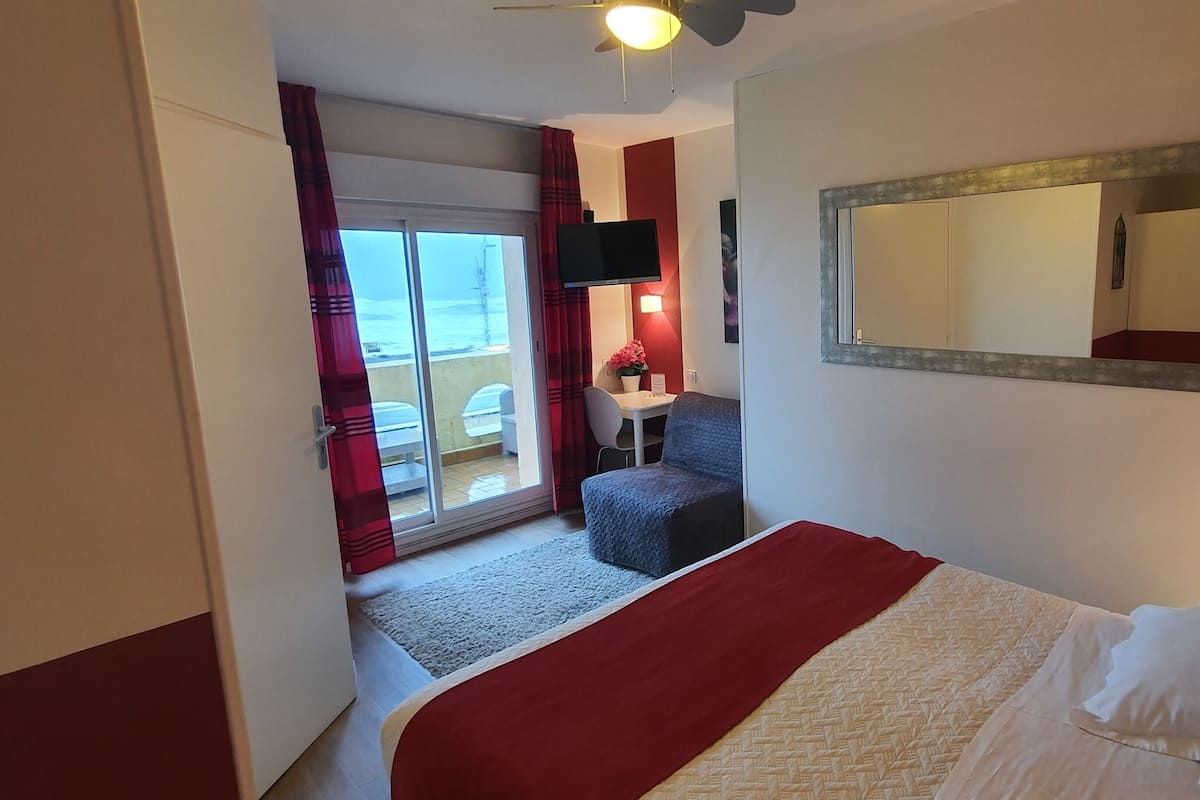 Habitación de hotel con cama, colcha a rayas rojas y blancas, espejo grande, televisor de pantalla plana, mesita con silla y vista al océano a través de una puerta corrediza.