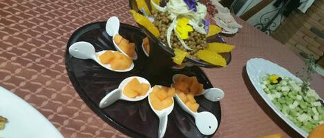 A table with a patterned tablecloth, a black tray with white serving spoons, and various food items including melon, grapes, and a salad.