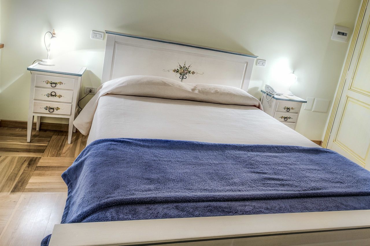 Ein ordentlich bezogenes Bett mit blauer Decke, ein Nachttisch mit Lampe und eine Wandleuchte.