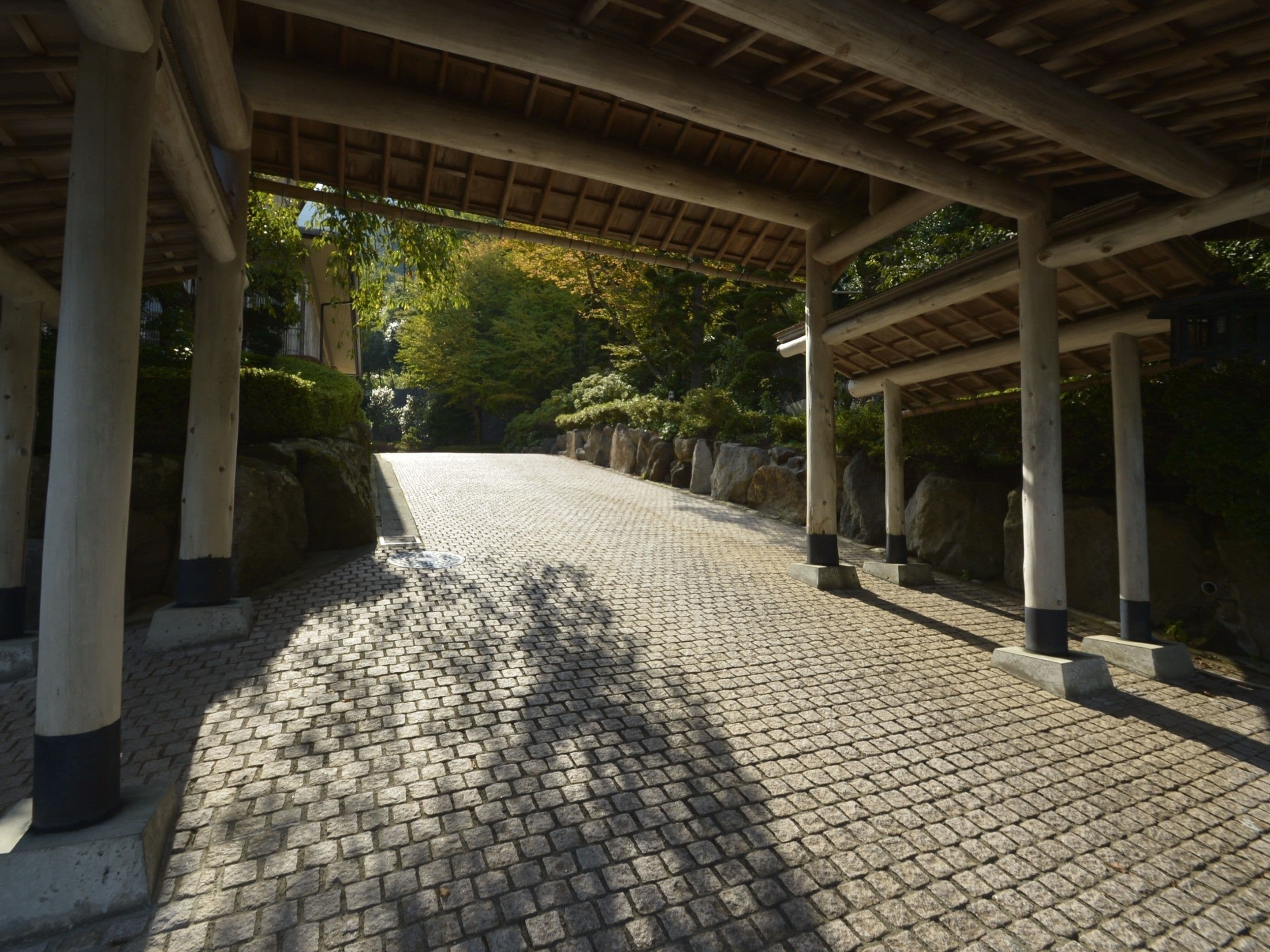 Une allée couverte dallée de pierres, avec des piliers en bois et un décor de fond composé d’arbres et de rochers.