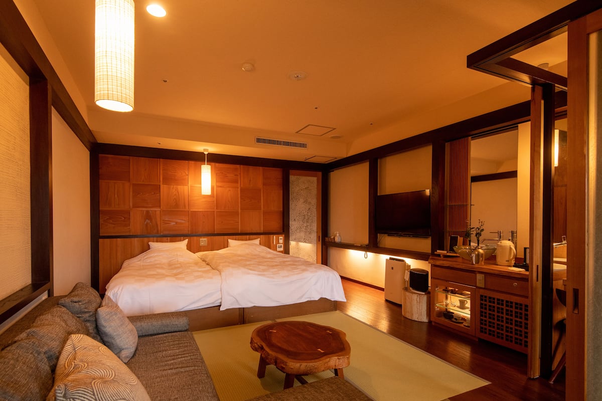 A hotel room with a large bed, a sofa, a small wooden stool, a television, and a bathroom with a sink and mirror.