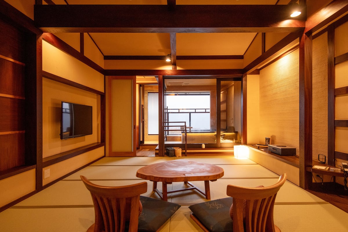 A traditional Japanese room with tatami flooring, a low table, two wooden chairs, a television, and a sliding door leading to another room.