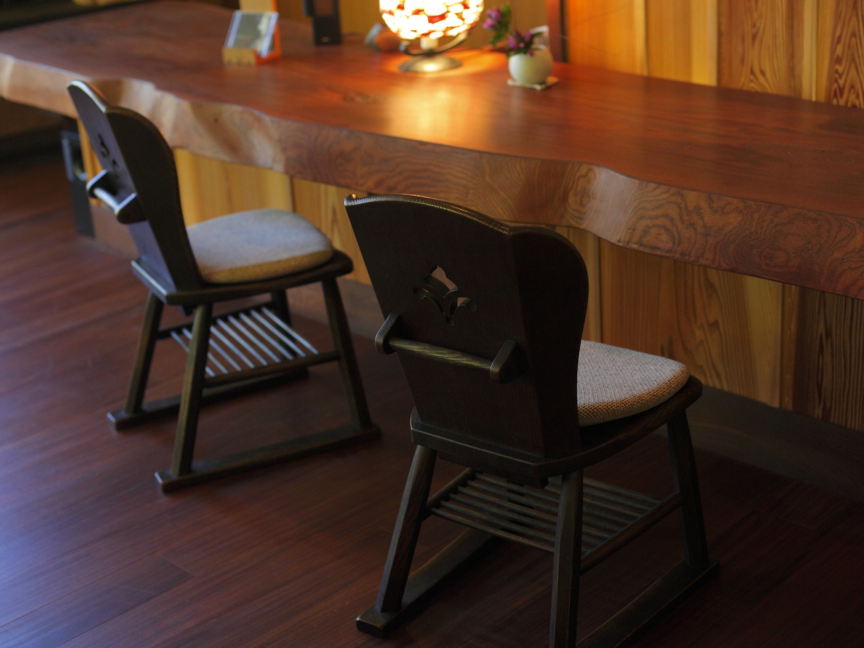 Dos sillas de madera con asientos acolchados y patas de metal, ubicadas frente a una barra de madera con una lámpara colgante.