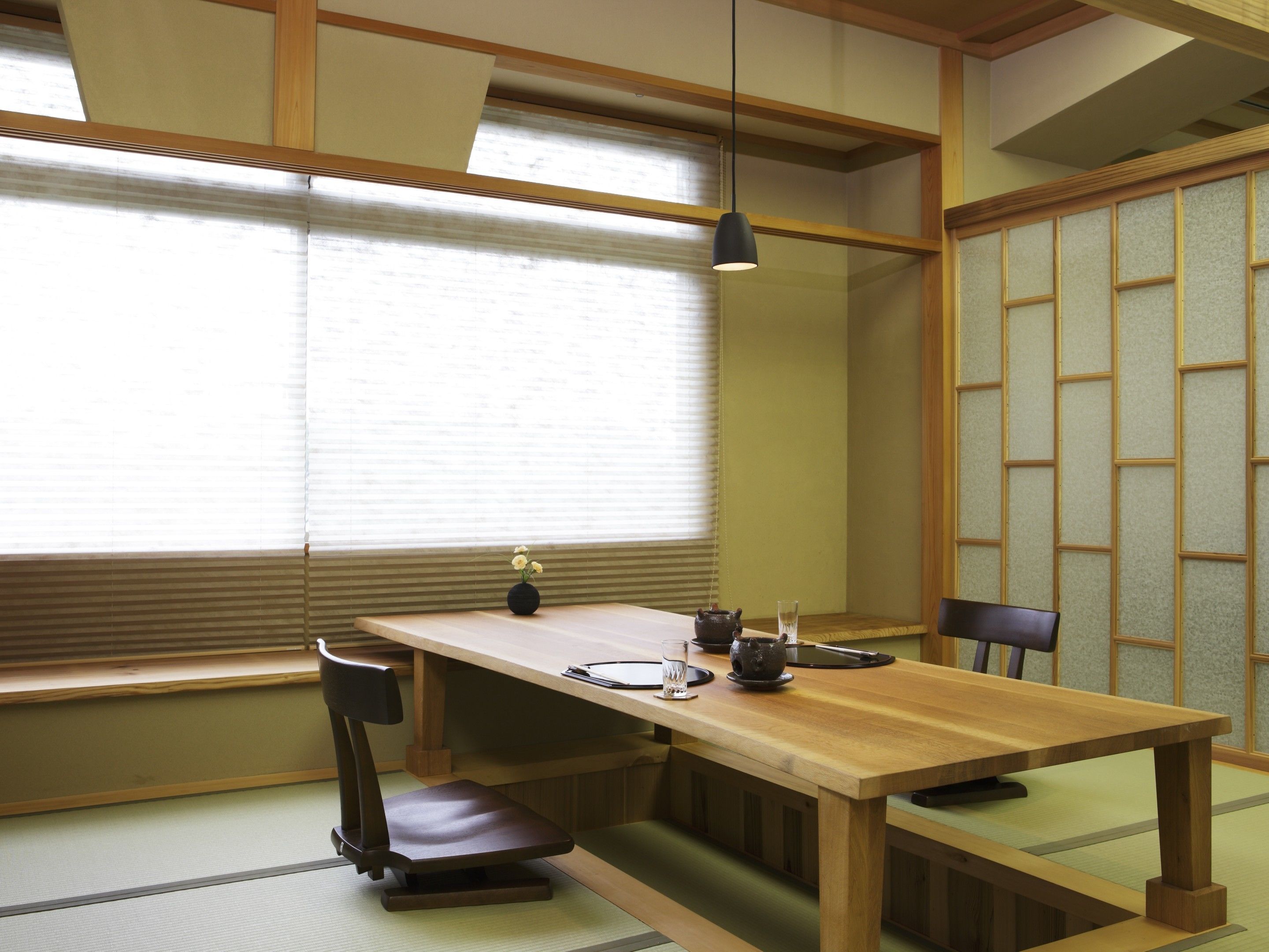 A traditional Japanese room with tatami flooring, a low wooden table, a black chair, a small vase with flowers, a teapot, and a glass of water.