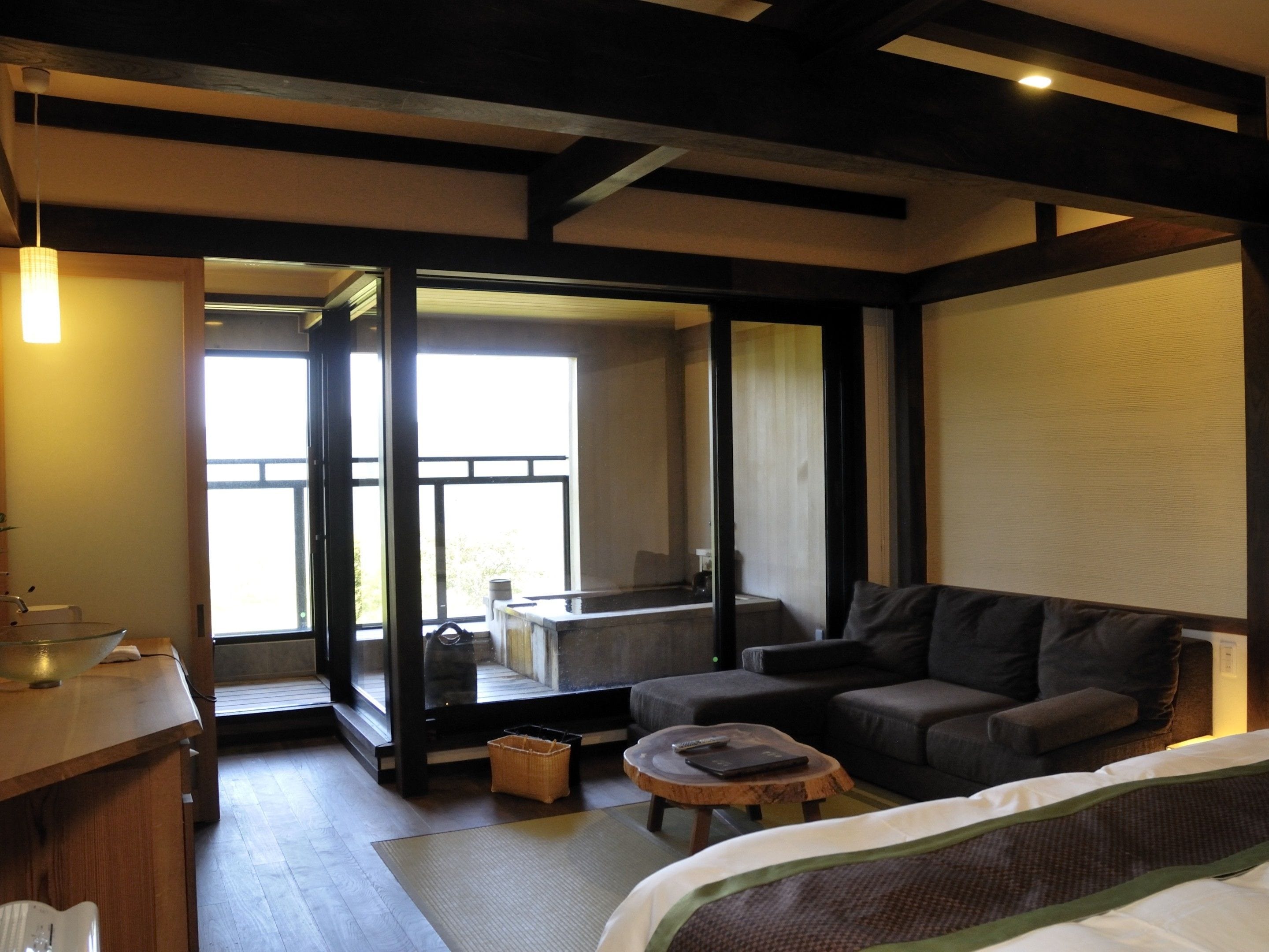 A modern hotel room with a sofa, a small table, a bed, and a large window offering a view of the outdoors.