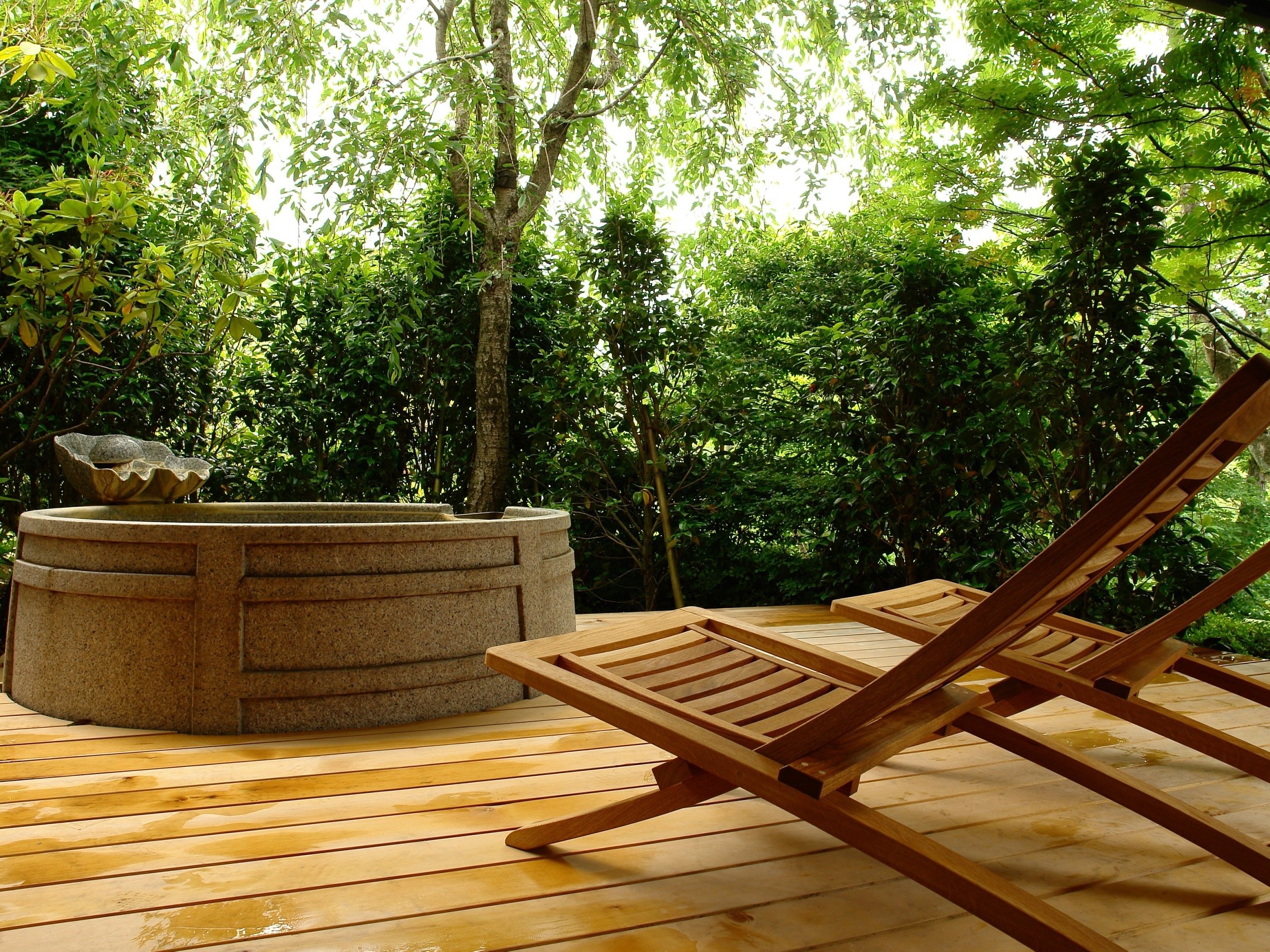 Una terraza de madera con un jacuzzi y sillones de descanso, rodeada de abundante vegetación.