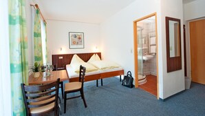 Une chambre d’hôtel avec un lit, un bureau et une chaise, une petite table avec une plante, et une salle de bain visible à travers une porte ouverte.
