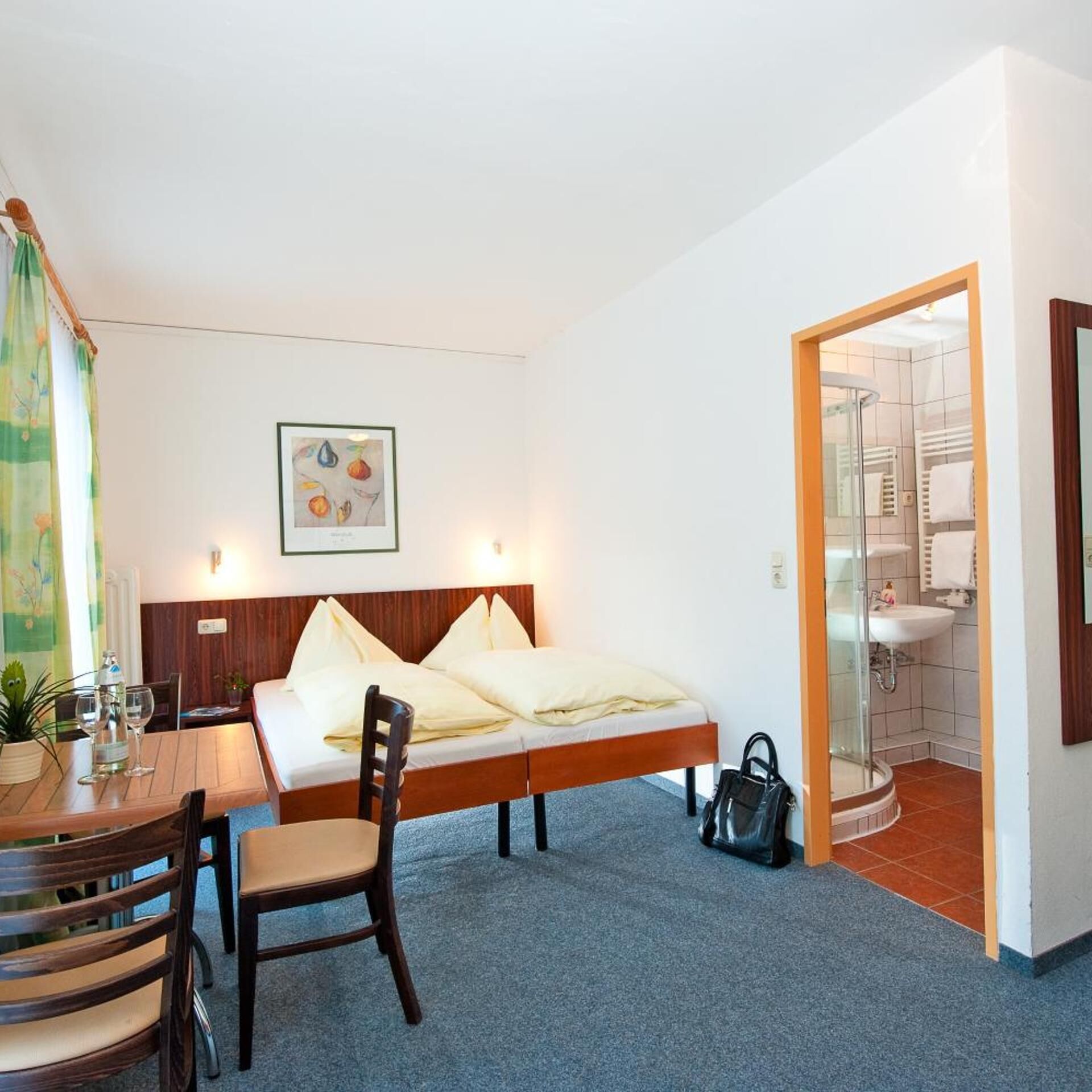 Ein Schlafzimmer mit Bett, Schreibtisch mit Stühlen, Badezimmer mit Waschbecken und Dusche sowie einem Bild an der Wand.