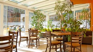 Ein helles Esszimmer mit Holztischen und -stühlen, großen Fenstern und Zimmerpflanzen.