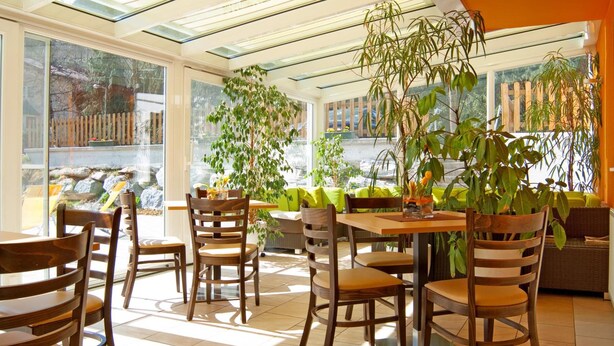 Un espace repas lumineux, avec des chaises et des tables en bois, de grandes fenêtres et des plantes d’intérieur.