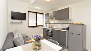 Une cuisine compacte avec des meubles gris, un four encastré, un évier et un réfrigérateur. On y trouve également un coin repas avec une table et des chaises, ainsi qu’un téléviseur à écran plat fixé au mur.