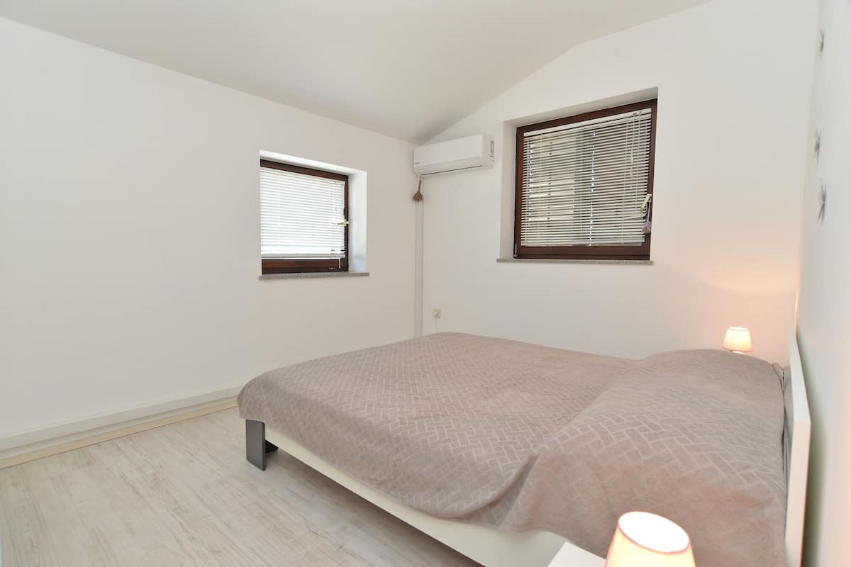 Een kleine kamer met een enkel bed en twee ramen, elk met jaloezieën, en een wandairconditioner.