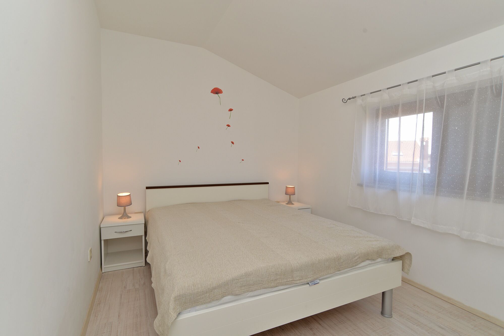 A bedroom with a bed, two bedside tables, a window with sheer curtains, and a wall decoration of red leaves.