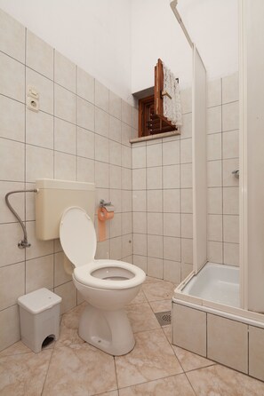 A bathroom with a toilet, a small trash bin, a shower area, and a tiled floor. - Apartment Milorad 324 (Medulin)