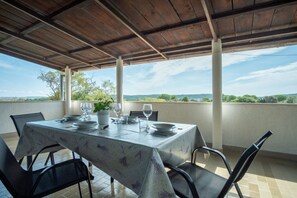 An outdoor dining area with a table set for four, surrounded by chairs, under a wooden pergola with a view of the countryside.