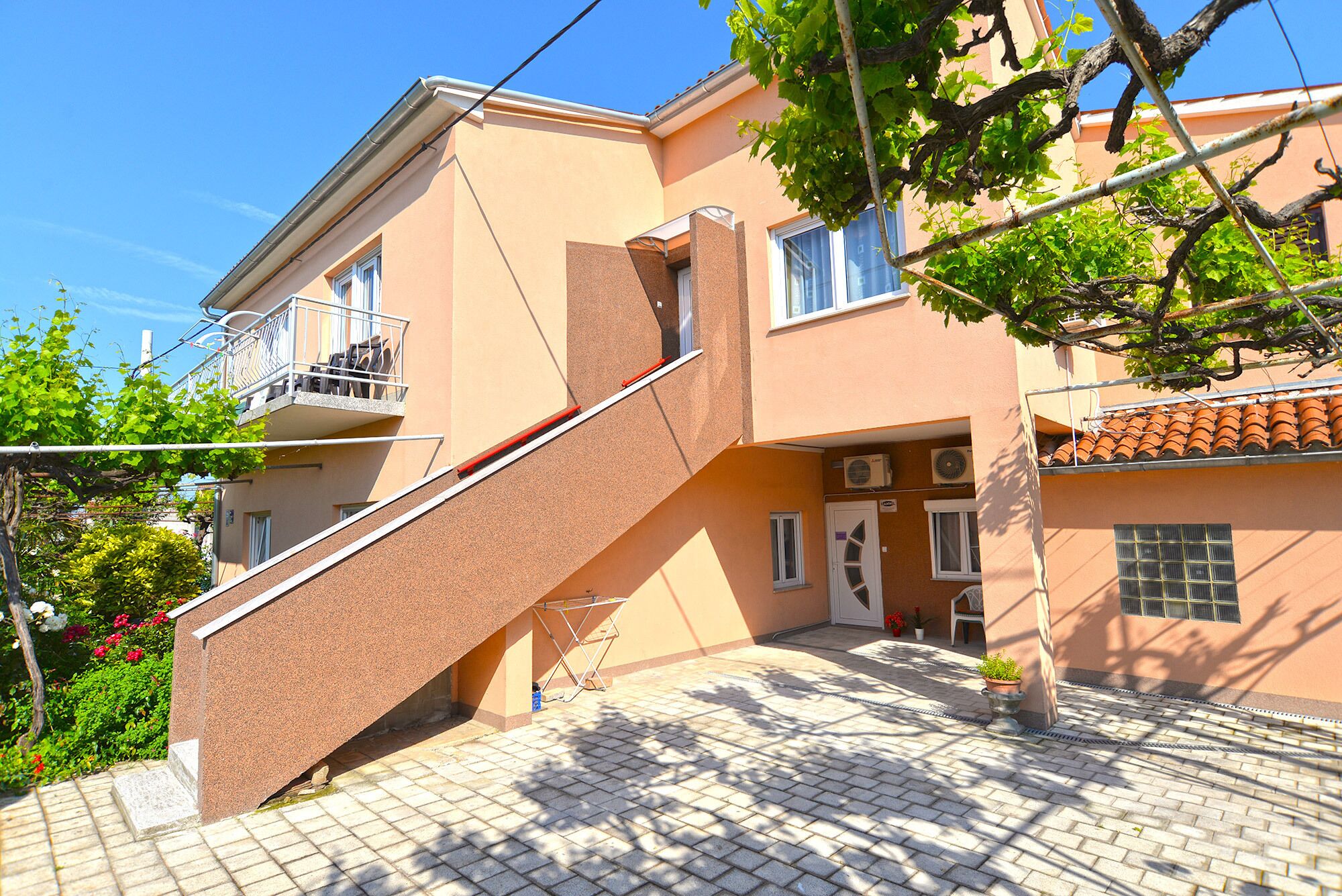 Um edifício de dois andares com escadaria que leva ao andar superior, uma varanda com mobiliário externo e um espaço de pátio com azulejos.